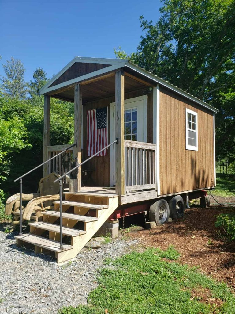 Small wood-sided manufactured home with front porch, stairs, and American flag, set among trees on a sunny day.