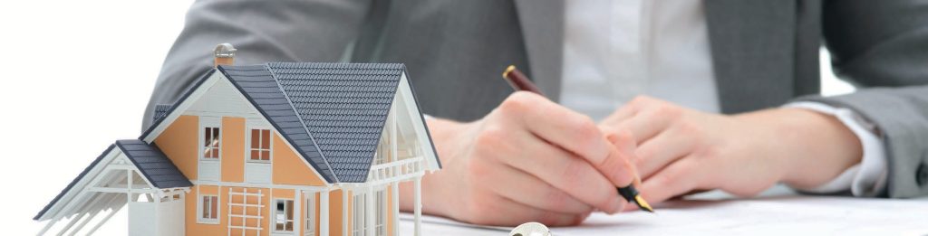 Small model house on a desk beside a person signing paperwork with a pen.