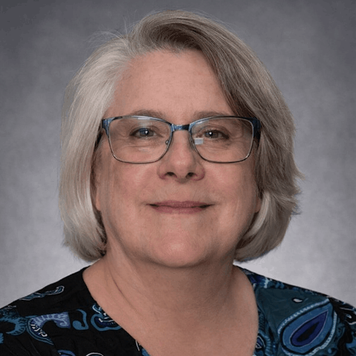 Professional headshot of a woman with short gray hair and glasses smiling against a neutral studio background.