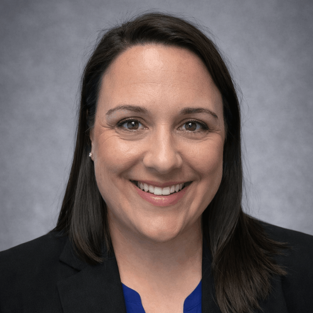 Professional headshot of a smiling woman with dark brown hair wearing a black blazer and blue top against a gray studio background.